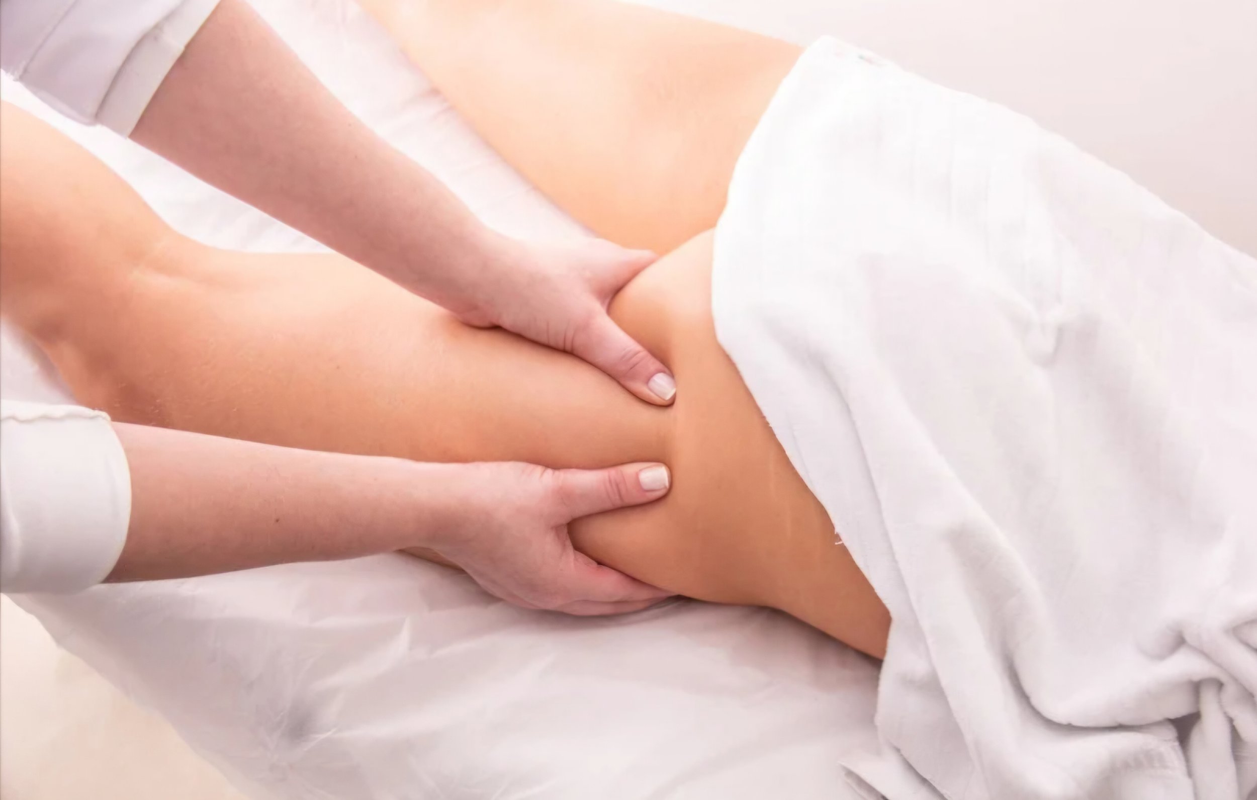 Person receiving lypossage massage on their thigh while lying on a massage table, draped with a white sheet. Moderate to deep pressure. Can add stones, cupping, aromatherapy, redlight to massage for ultimate relaxation, serenity, and massage spa