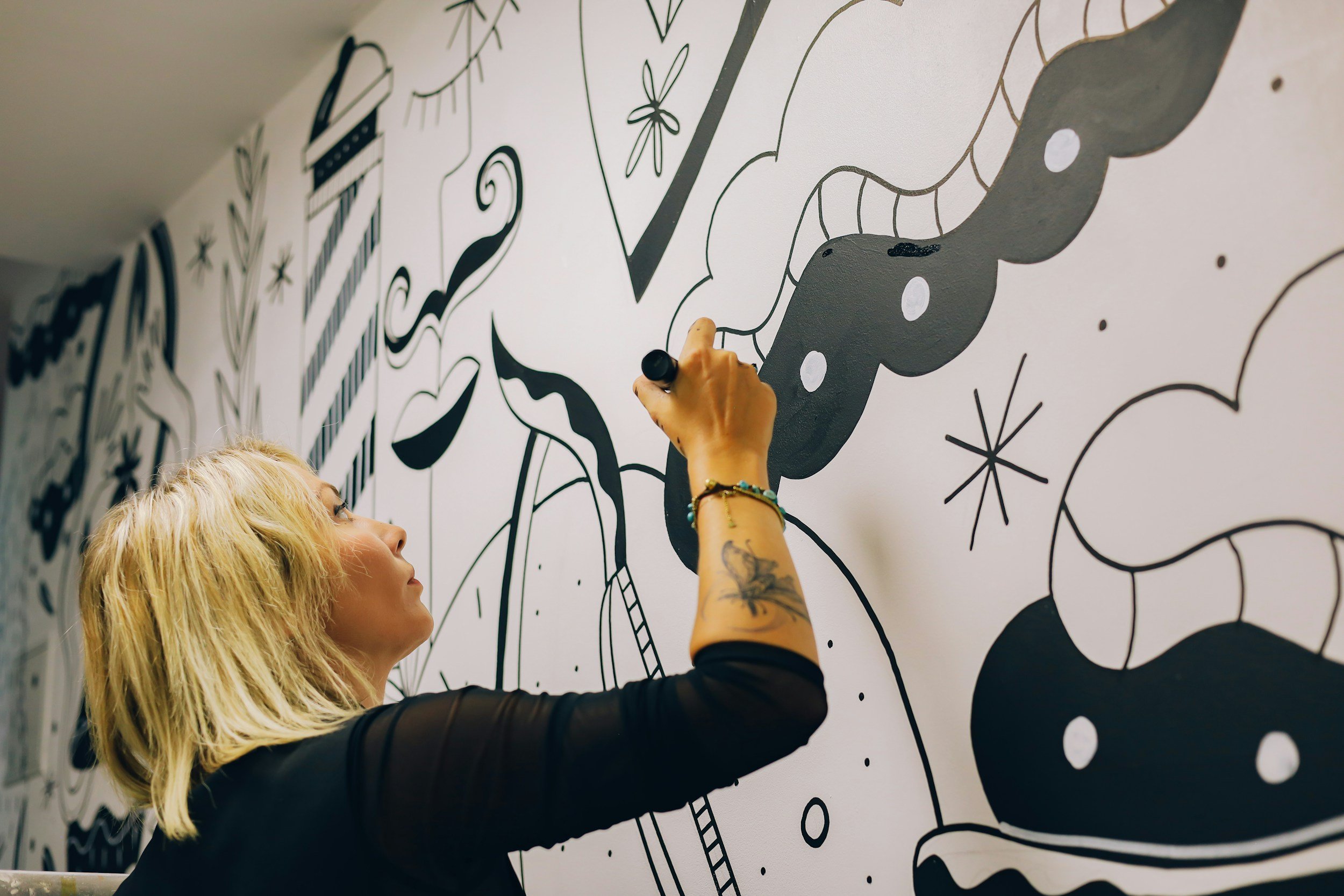 A woman with blonde hair drawing with black marker on a large abstract black and white mural wall.