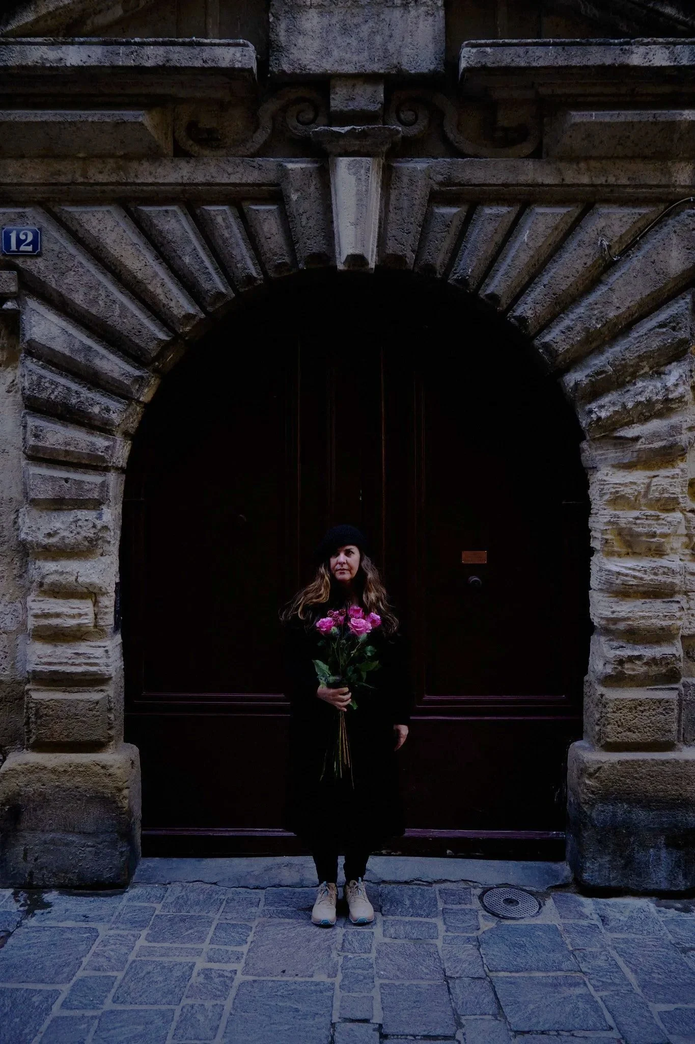 A woman with long hair wearing a black coat and a black beanie holding a bouquet of pink flowers, standing in front of a dark wooden arched door in a stone building.