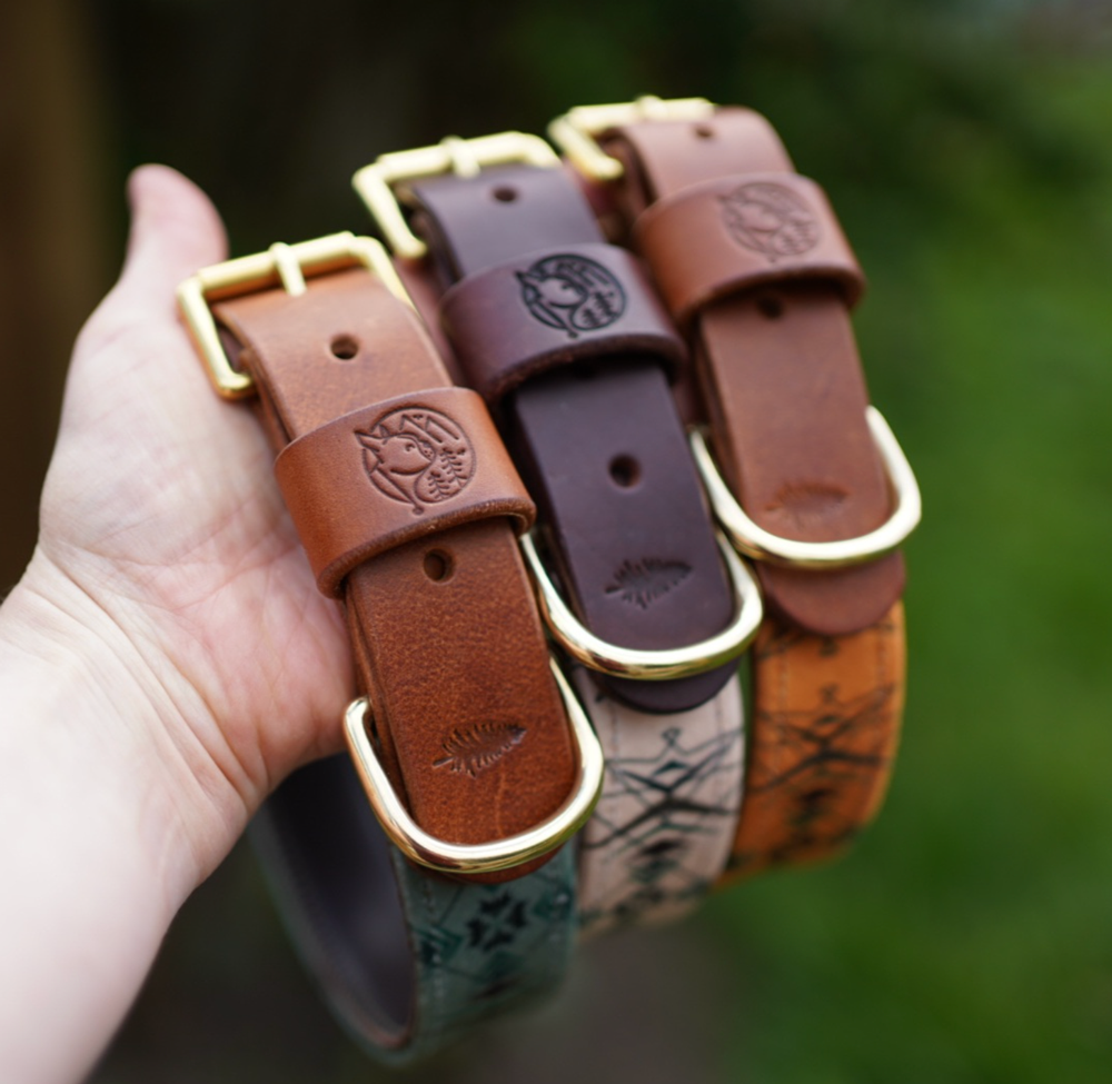 A hand holding three leather dog collars with brass buckles, each featuring a carved fox and fish design, in an outdoor setting with blurred greenery in the background.