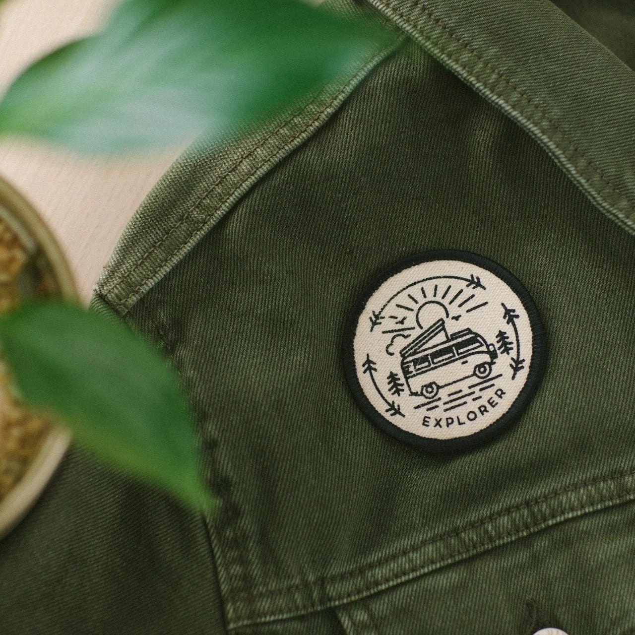 Close-up of a green fabric backpack with a circular embroidered patch featuring a van, trees, sun rays, and the word 'EXPLORE'.
