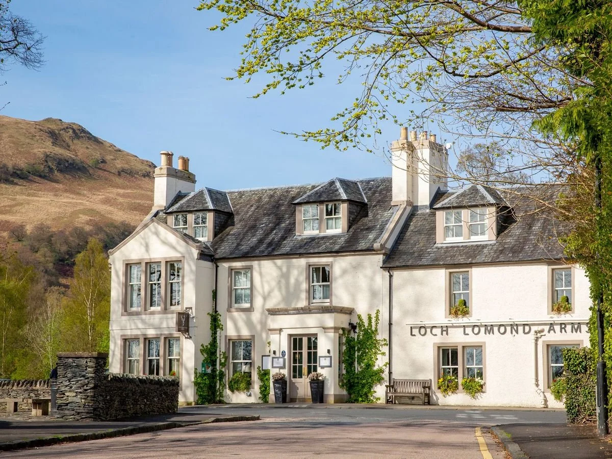 A white multi-story building with a sign reading 'Loch Lomond Arms' and surrounded by trees, with mountains in the background.