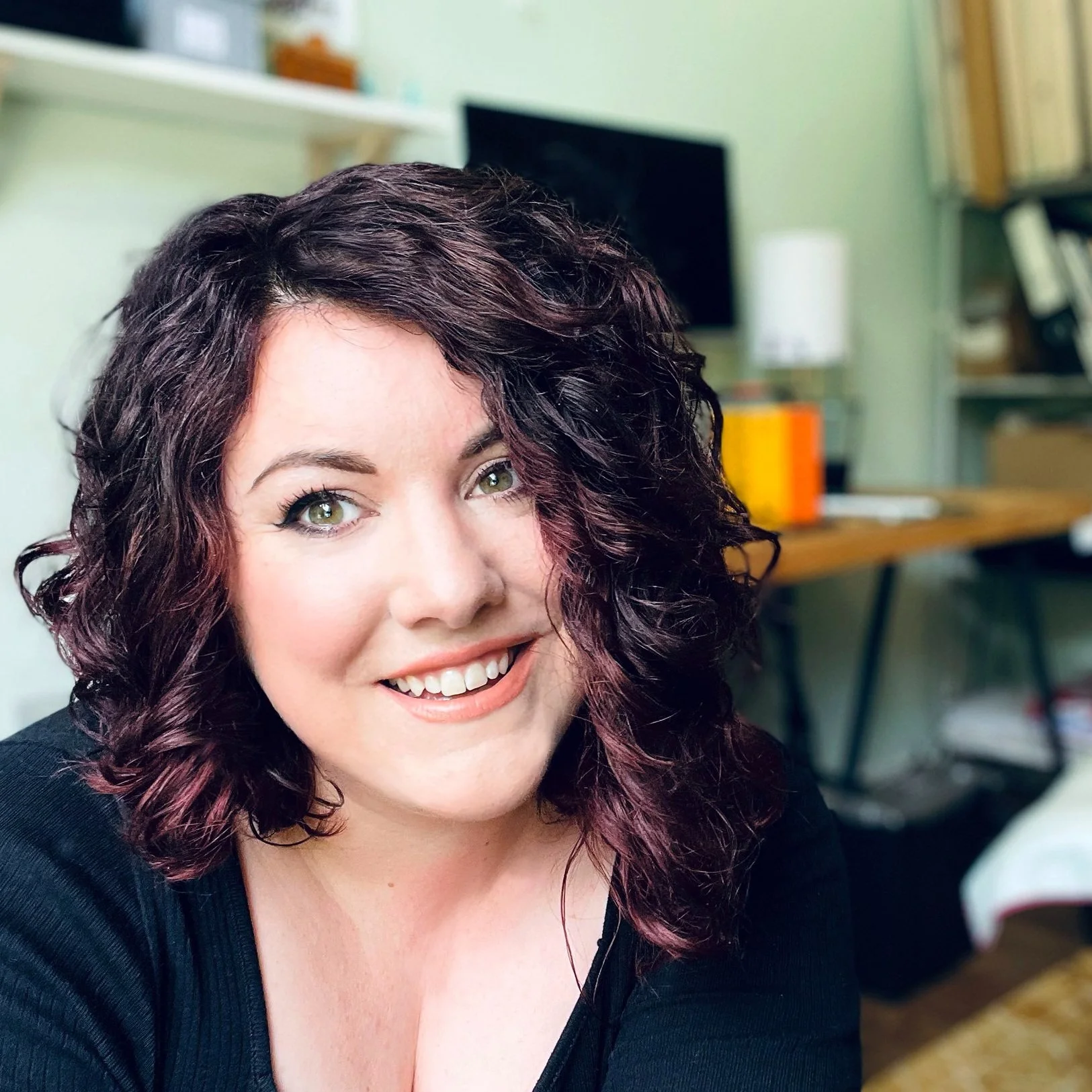 A woman with curly dark hair and green eyes smiling in an indoor setting, with bookshelves, a computer monitor, and a lamp in the background.