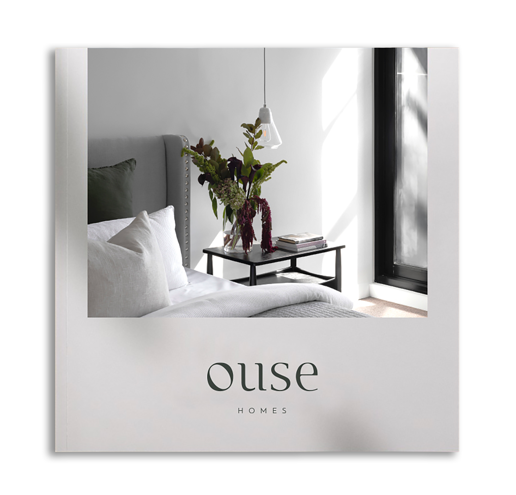 Close-up of a white book cover with the text 'our home' and an image of a bedroom interior, featuring a bed with gray headboard and pillows, a side table with a vase of flowers, and a window with blinds.
