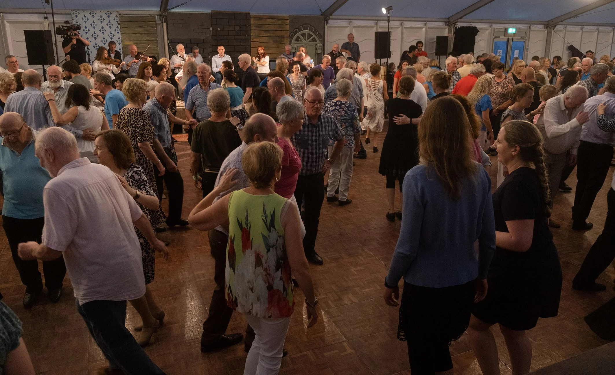 People dancing and socializing in a large indoor event tent with musicians playing on a stage in the background.