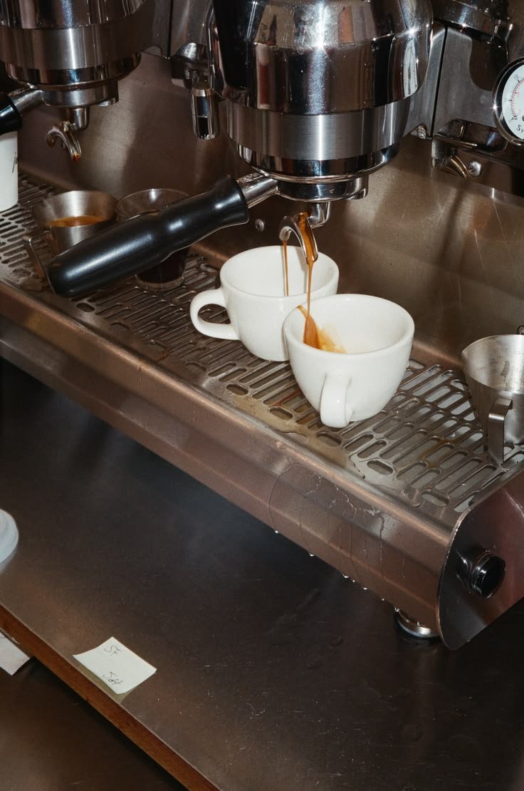 Coffee service for two: white coffee cups on a mug warmer with espresso being dispensed from an espresso machine into the cups in D F W, The Village Lounge.