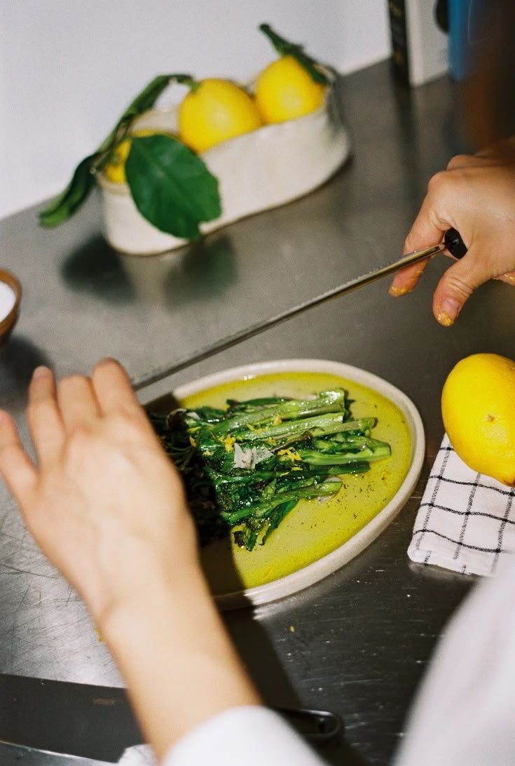Photograph of a cook garnishing a dish: zesting citrus over green vegetables, on a white plate with yellow oil, in a kitchen setting with fresh yellow lemons in the background.