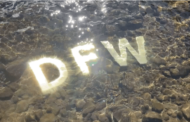 Solid brass signage letters D, F, and W sitting on a rocky river bottom, under flowing water with sunlight reflections.