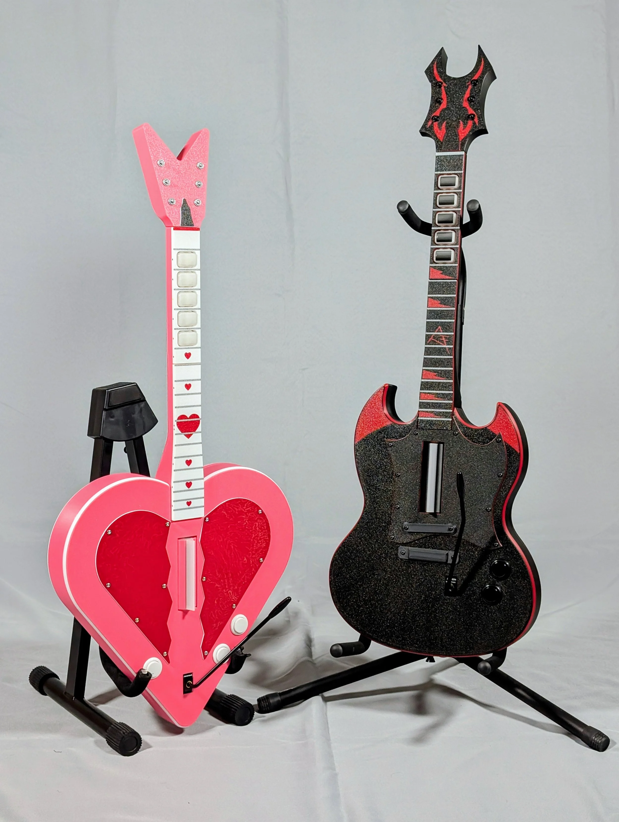 Two electric guitar-shaped objects on black stands, one pink with heart designs and the other black with red accents, placed on a gray surface against a gray background.
