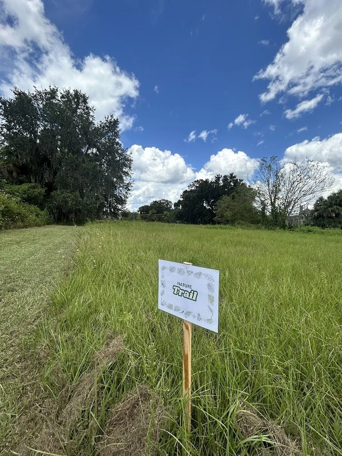 A grassy nature trail marked by a small white sign with decorative borders, situated in a lush green outdoor setting under a partly cloudy blue sky, with trees in the background.