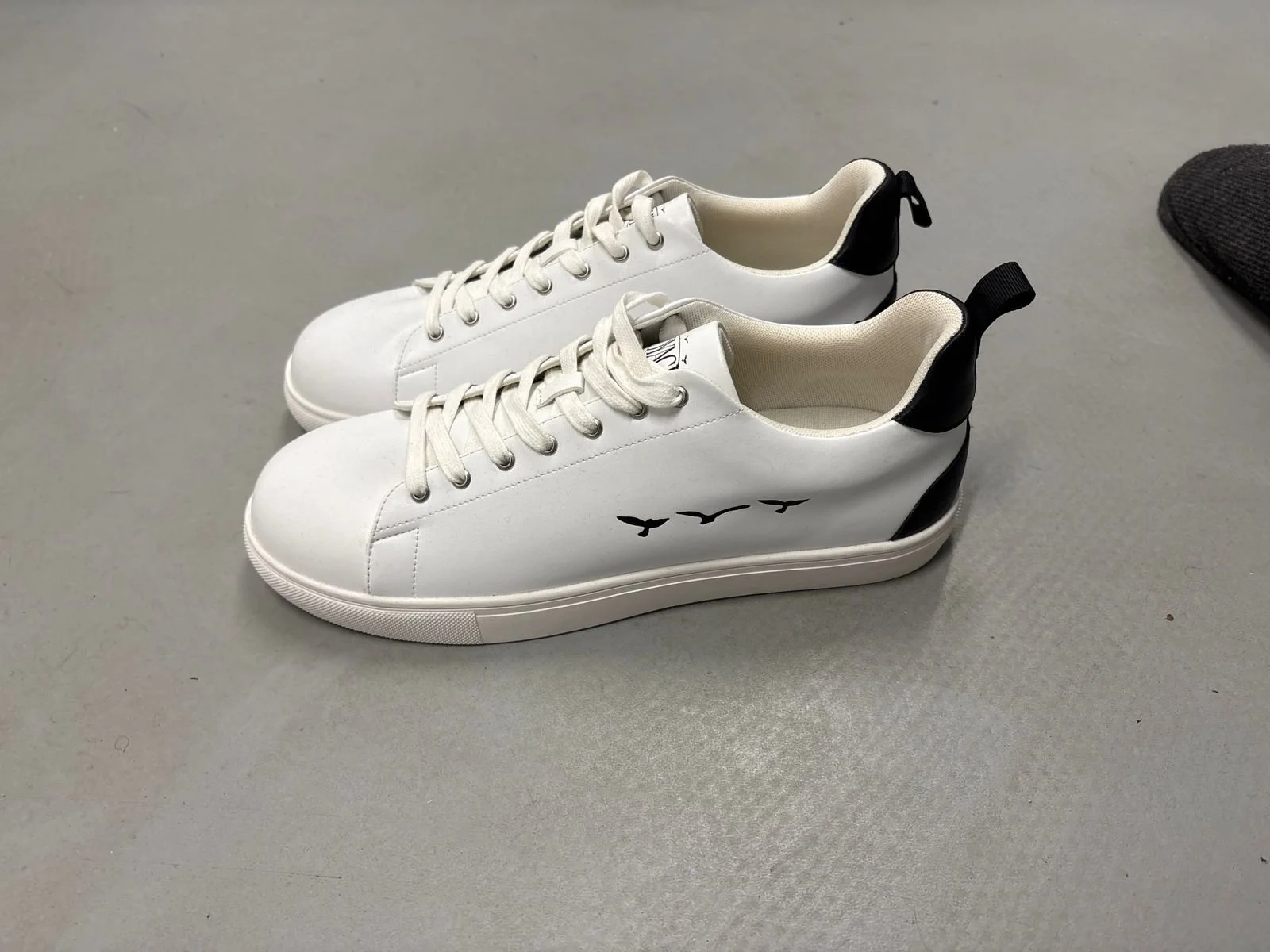 White sneakers with black heel accents and a bird silhouette logo on the side, placed on a light gray surface.