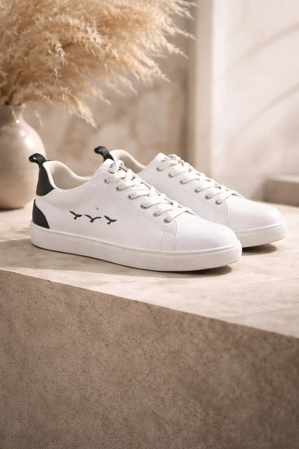 White sneakers with black accents and bird silhouettes on the side, placed on a beige surface with a vase of dried plants in the background.