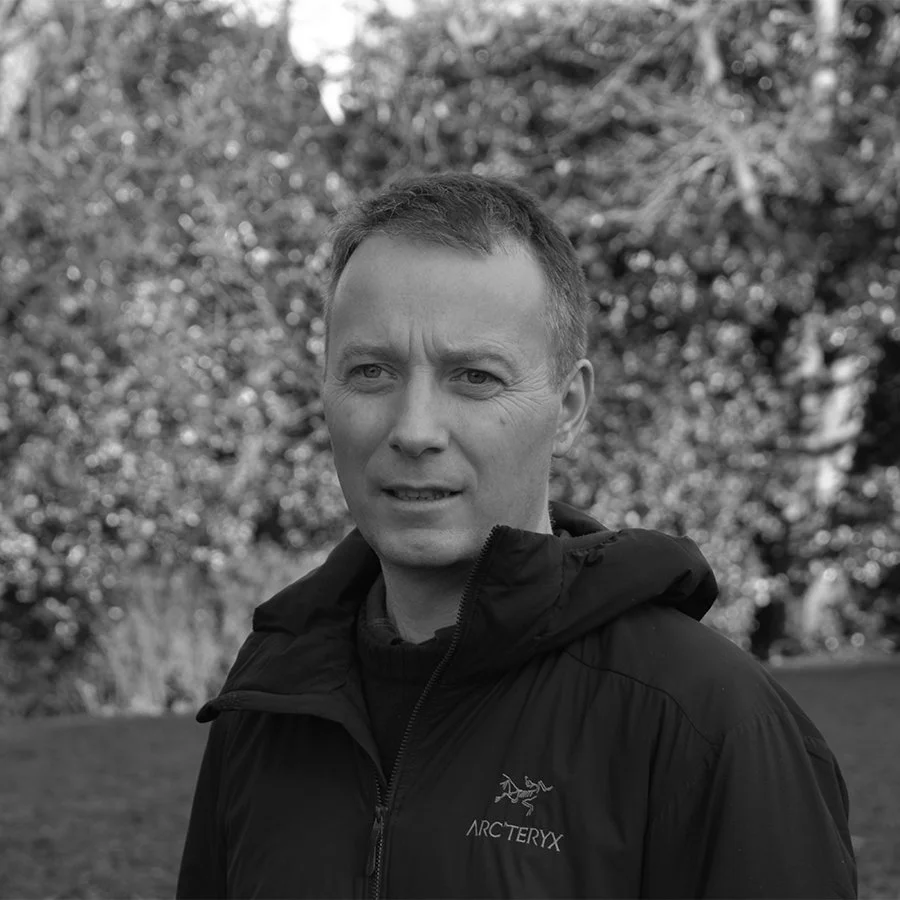 Black and white photo of a man outdoors wearing a jacket, with a background of trees and foliage.