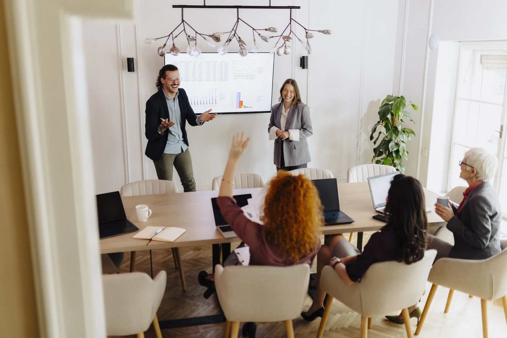 A business meeting in a modern conference room where a man is giving a presentation on a screen, with a woman standing next to him. Several women are seated around the table, one raising her hand. The room has a large window, a plant, and a stylish light fixture.