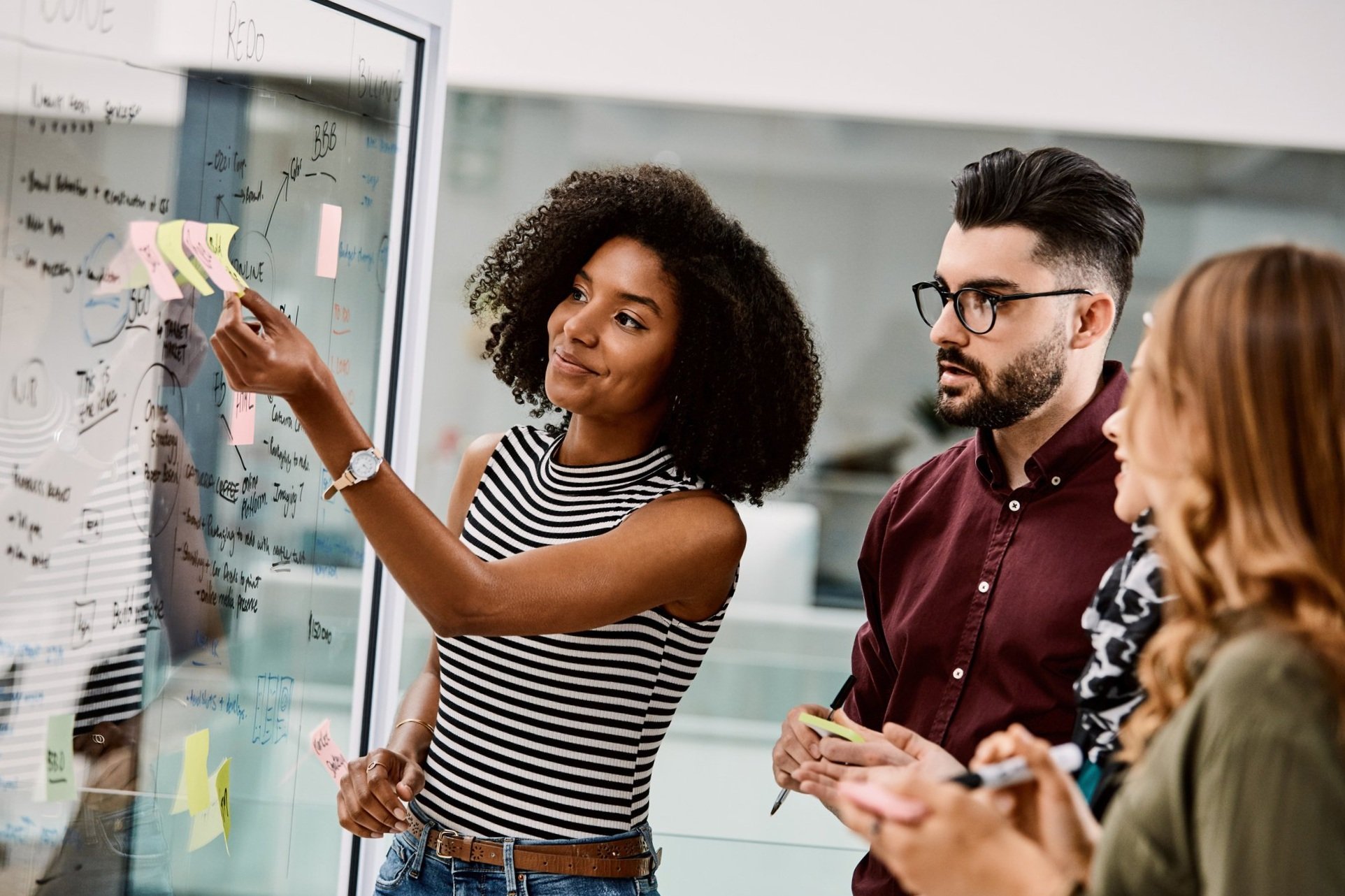 Three people in an office near a glass wall with notes and diagrams. One woman with curly hair in a striped sleeveless top is placing sticky notes on the glass wall, while a man with glasses and a beard in a maroon shirt and a woman with long reddish hair in a green jacket look on and take notes.