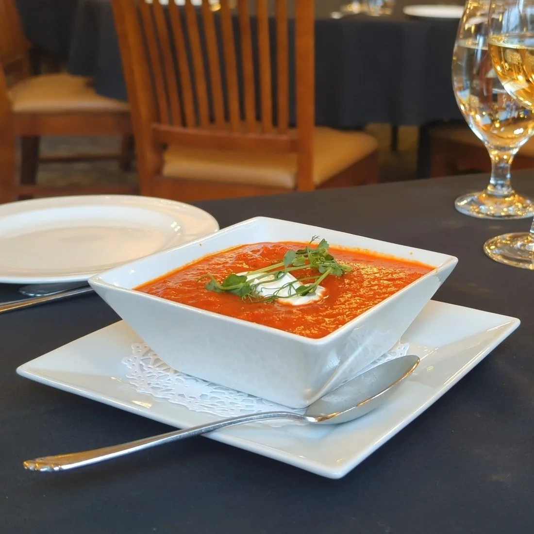 A bowl of tomato soup garnished with fresh herbs and a dollop of sour cream on a white plate with a spoon, on a black table in a restaurant setting.