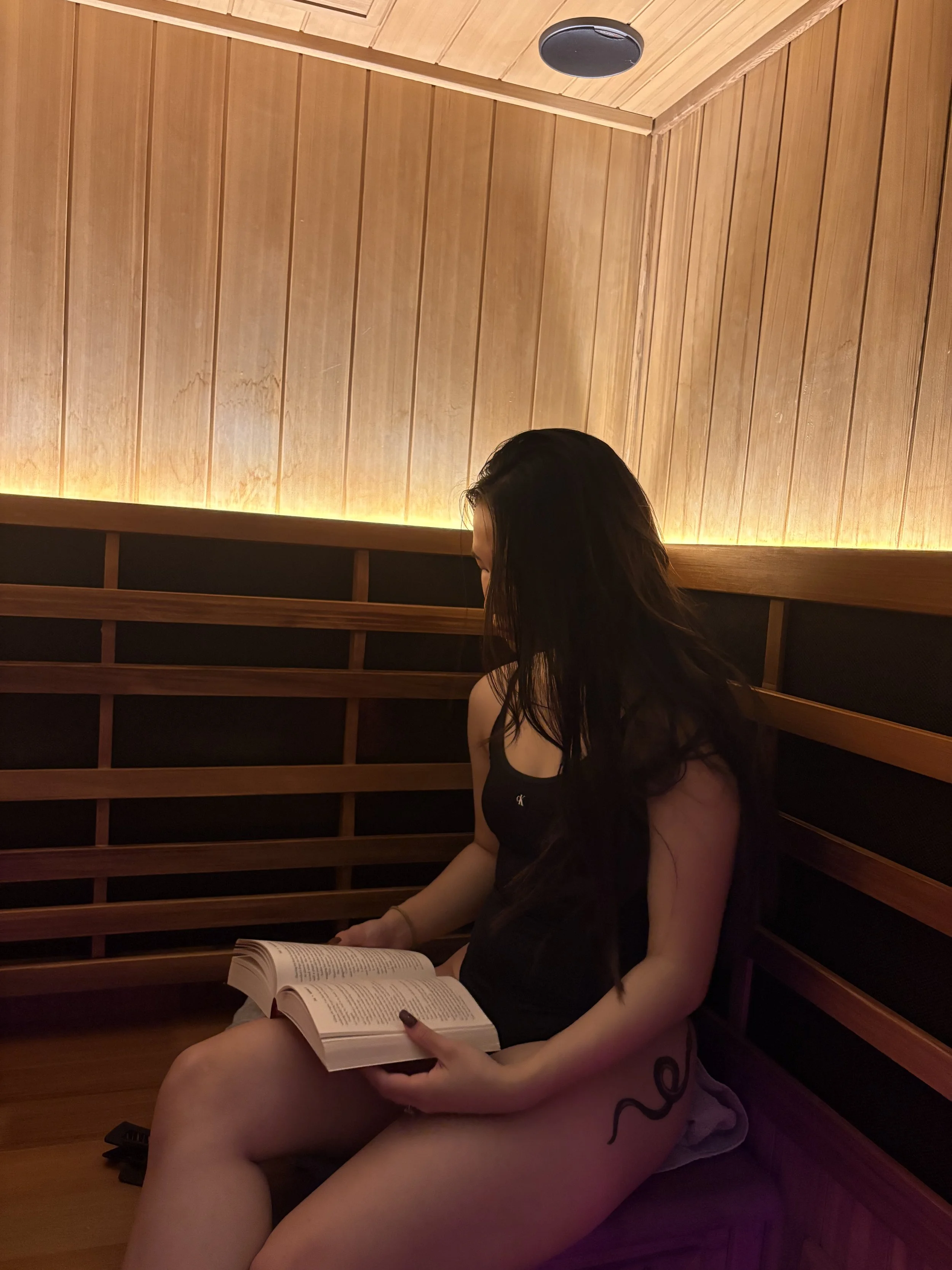 A woman with long dark hair sitting on a wooden bench, reading a book in a cozy room with warm wooden paneling and soft lighting.