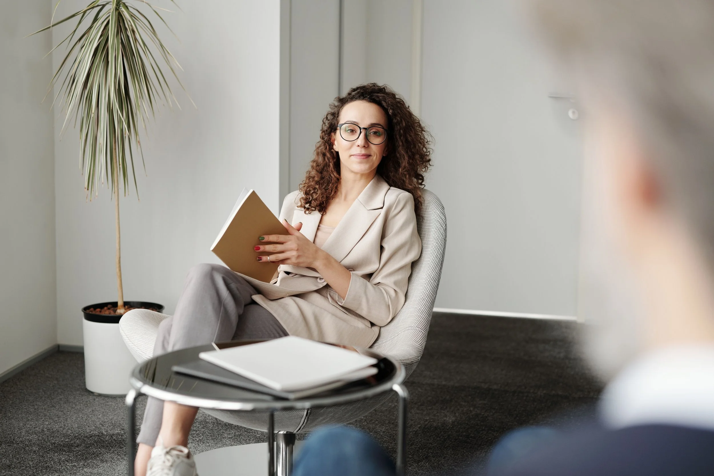 A woman with curly hair, glasses, and wearing a light blazer, sitting in an office chair, holding a notebook, with a potted plant and a small table in the background.