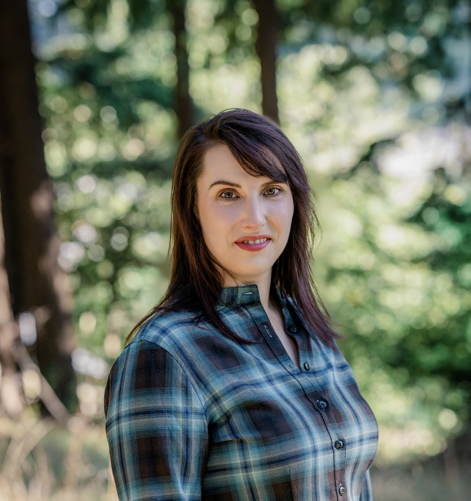 A woman with dark brown hair, wearing a blue plaid shirt, outdoors with a blurred lush green forest background.