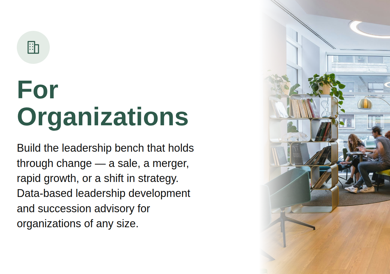Modern office space with large windows, bookshelf, plants, and people sitting on chairs, working on laptops.