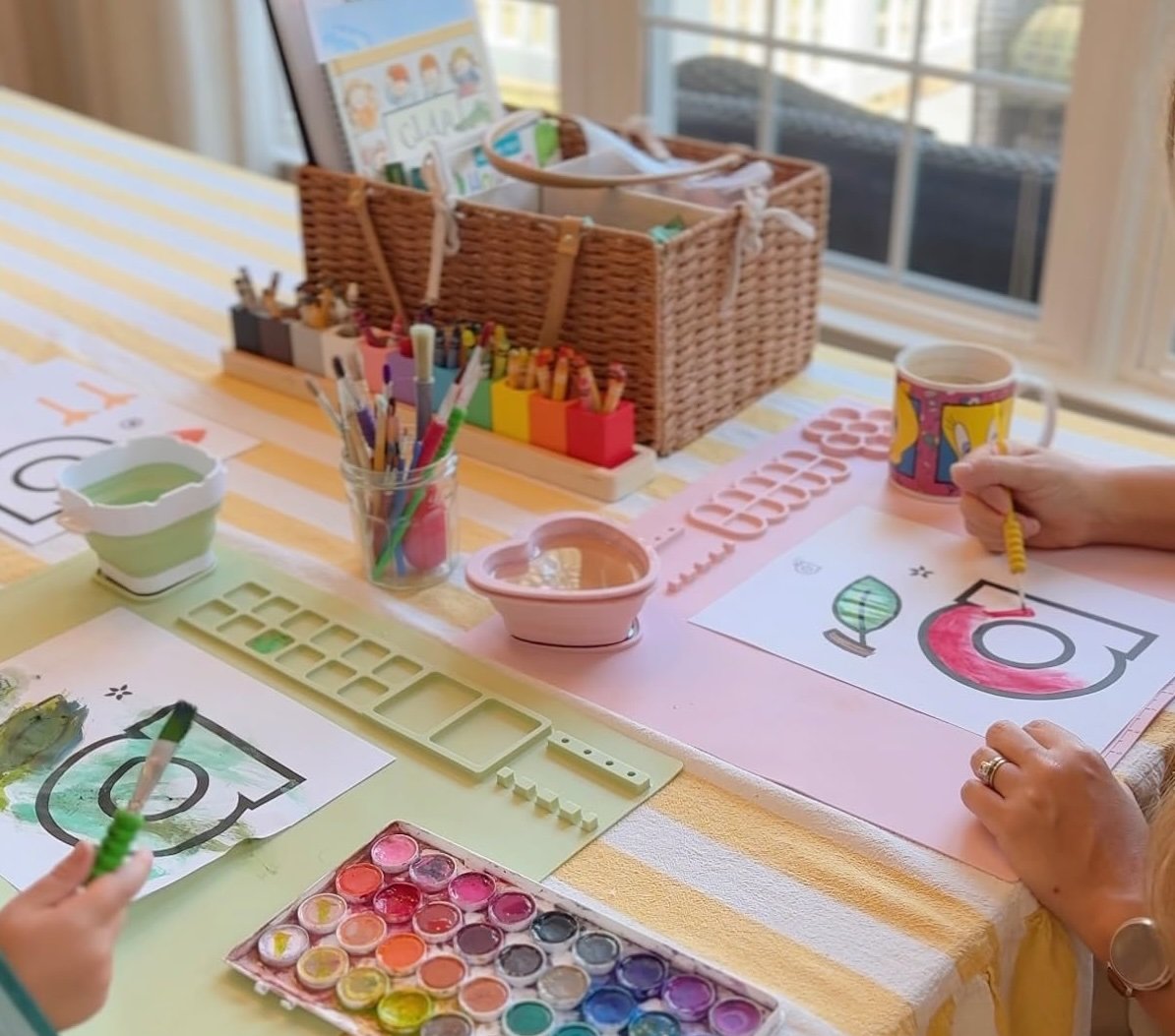 Morning basket on a striped kitchen table with watercolors, letter pages, a coffee mug and a woven basket of supplies
