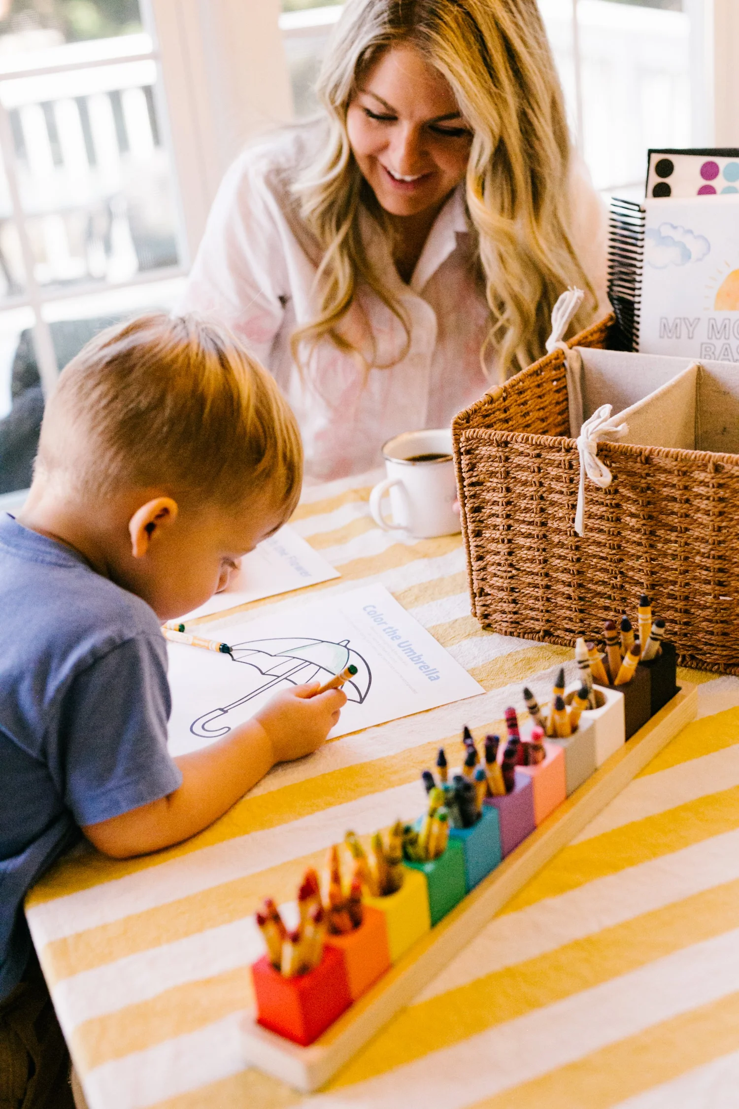 Toddler coloring a morning basket activity page at the kitchen table beside a woven basket and crayons, with mom smiling nearby
