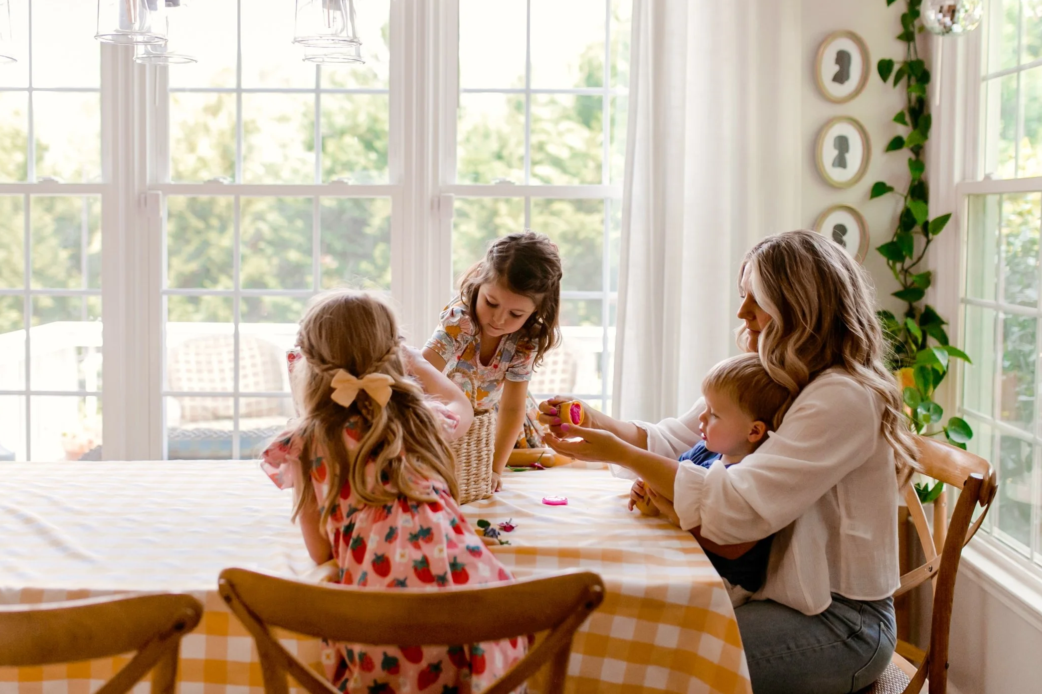 Mom and three kids playing with a morning basket at a gingham kitchen table.