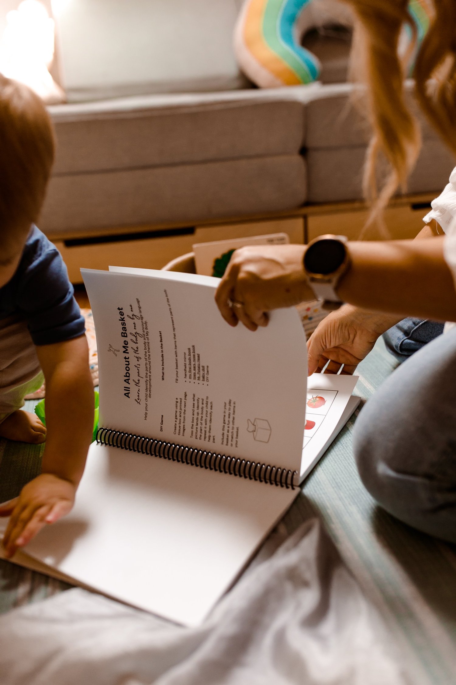 Toddler turning the pages of a spiral-bound activity book on the floor with mom