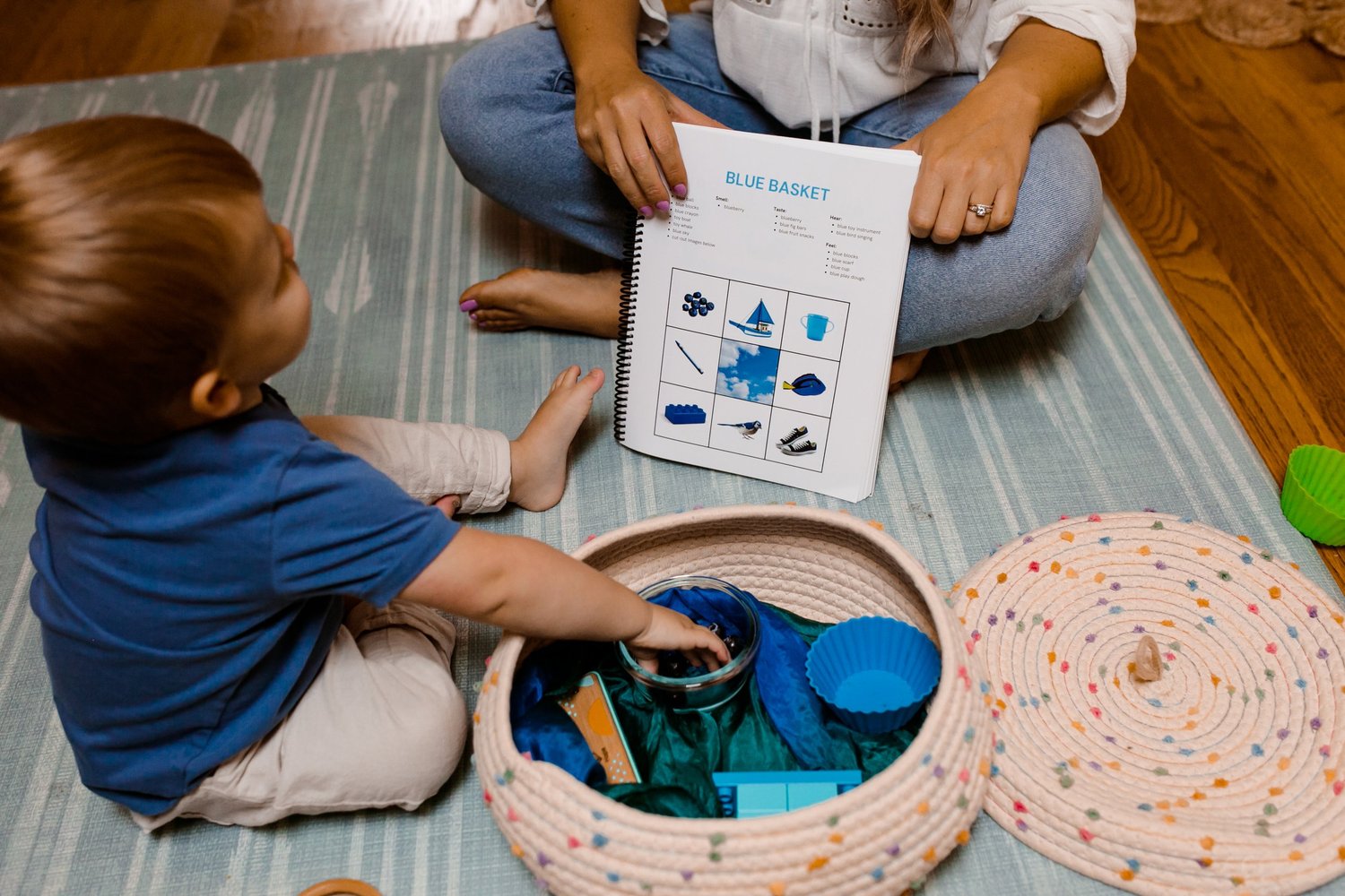 Toddler sitting on the floor exploring a basket of soft toys while mom sits nearby