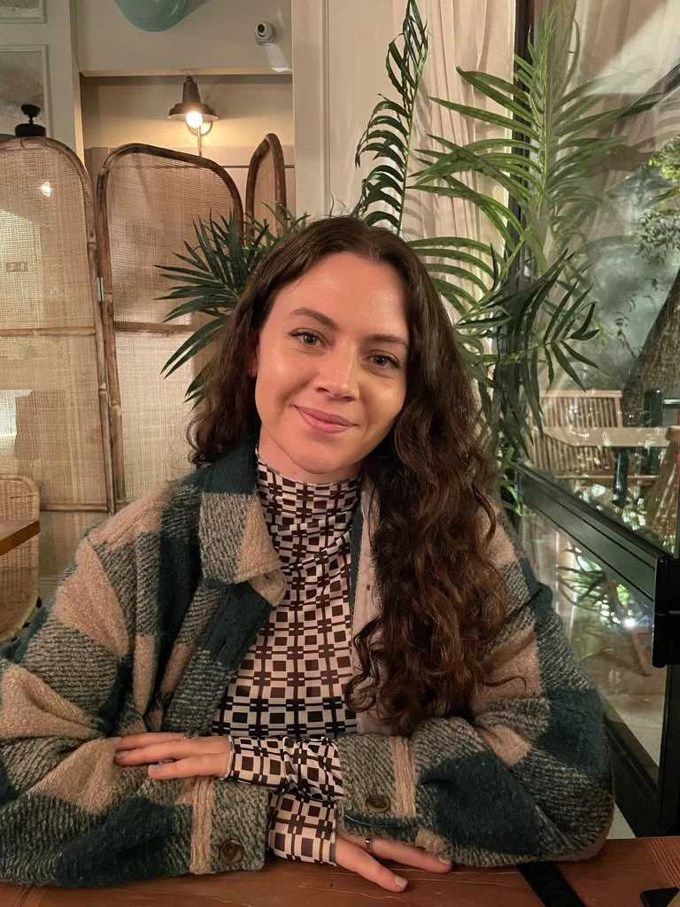 A woman with long curly hair sitting at a table in a cozy indoor setting with plants and warm lighting, wearing a plaid jacket over a geometric patterned top.