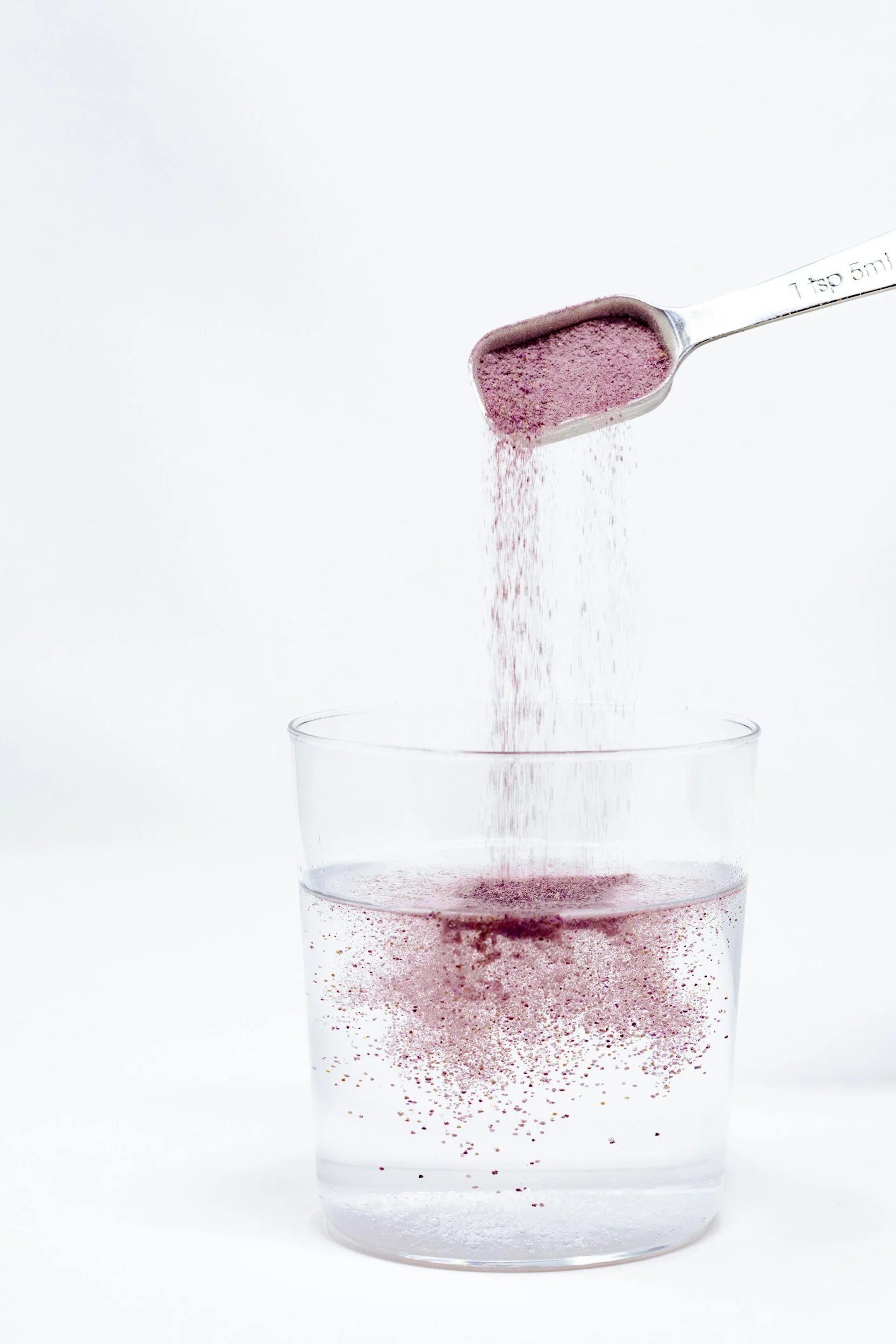 A metal teaspoon pouring pink unicorn-colored glitter into a clear glass of water, creating a sparkling effect.