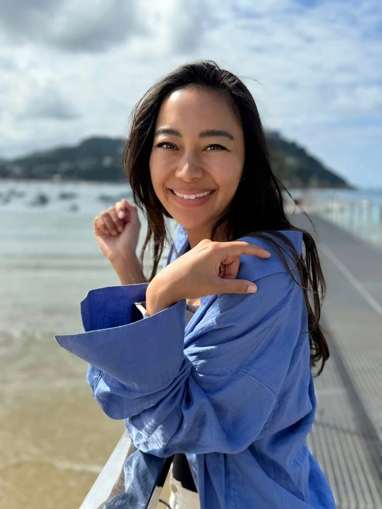 A woman with long dark hair smiling and making a playful fist gesture, standing on a seaside walkway with a mountain and cloudy sky in the background.