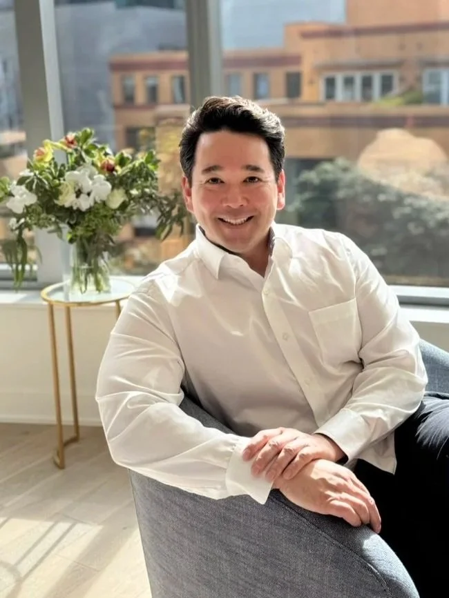 A man in a white dress shirt sitting in a modern room with large windows and a view of buildings outside, smiling at the camera. There is a vase of white flowers and greenery on a small table behind him.