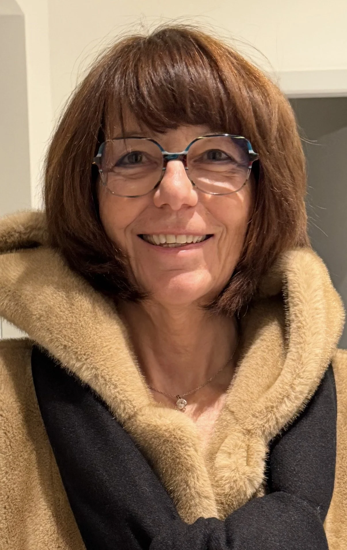 A woman with short reddish-brown hair and glasses, smiling, wearing a black and tan coat with a fuzzy collar, indoors.