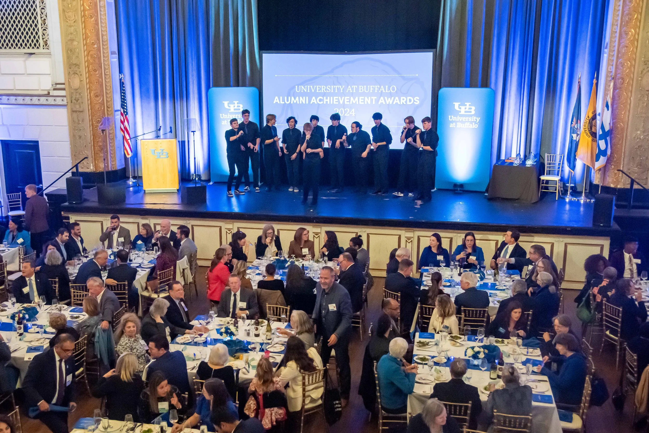 University at Buffalo Alumni Achievement Awards event with attendees seated at tables, and a stage with a group of people standing and speaking in front of a large screen.