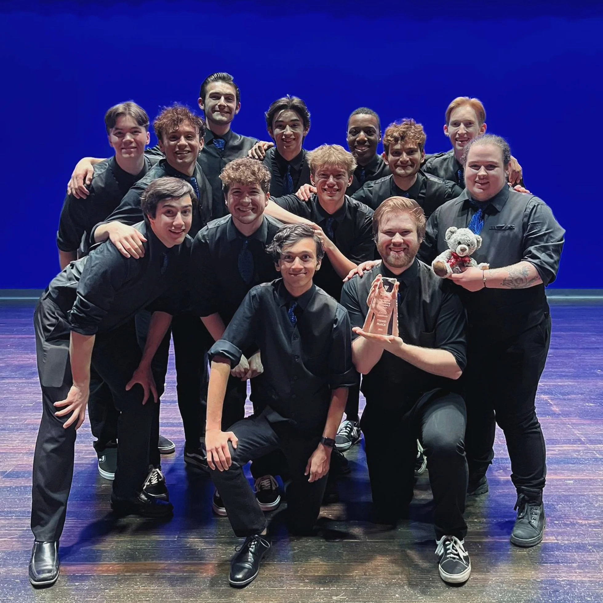 Group of young people wearing black shirts and pants, some with blue ties, posing on stage against a dark blue background, holding up a trophy and a small teddy bear.