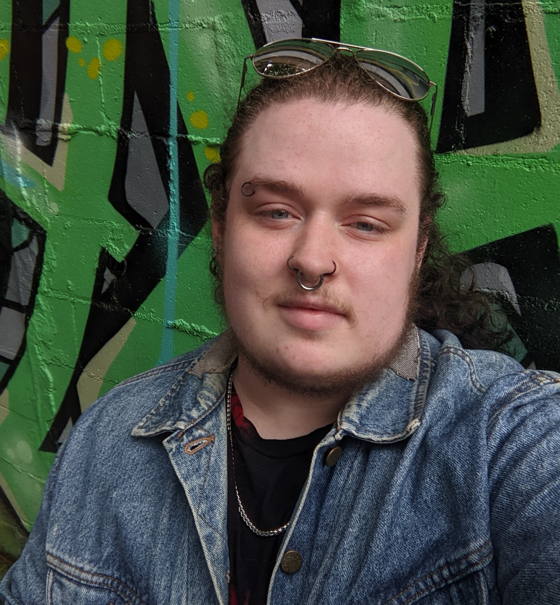 A young person with long curly hair, pierced nose, and sunglasses on head taking a selfie in front of a green graffiti wall.