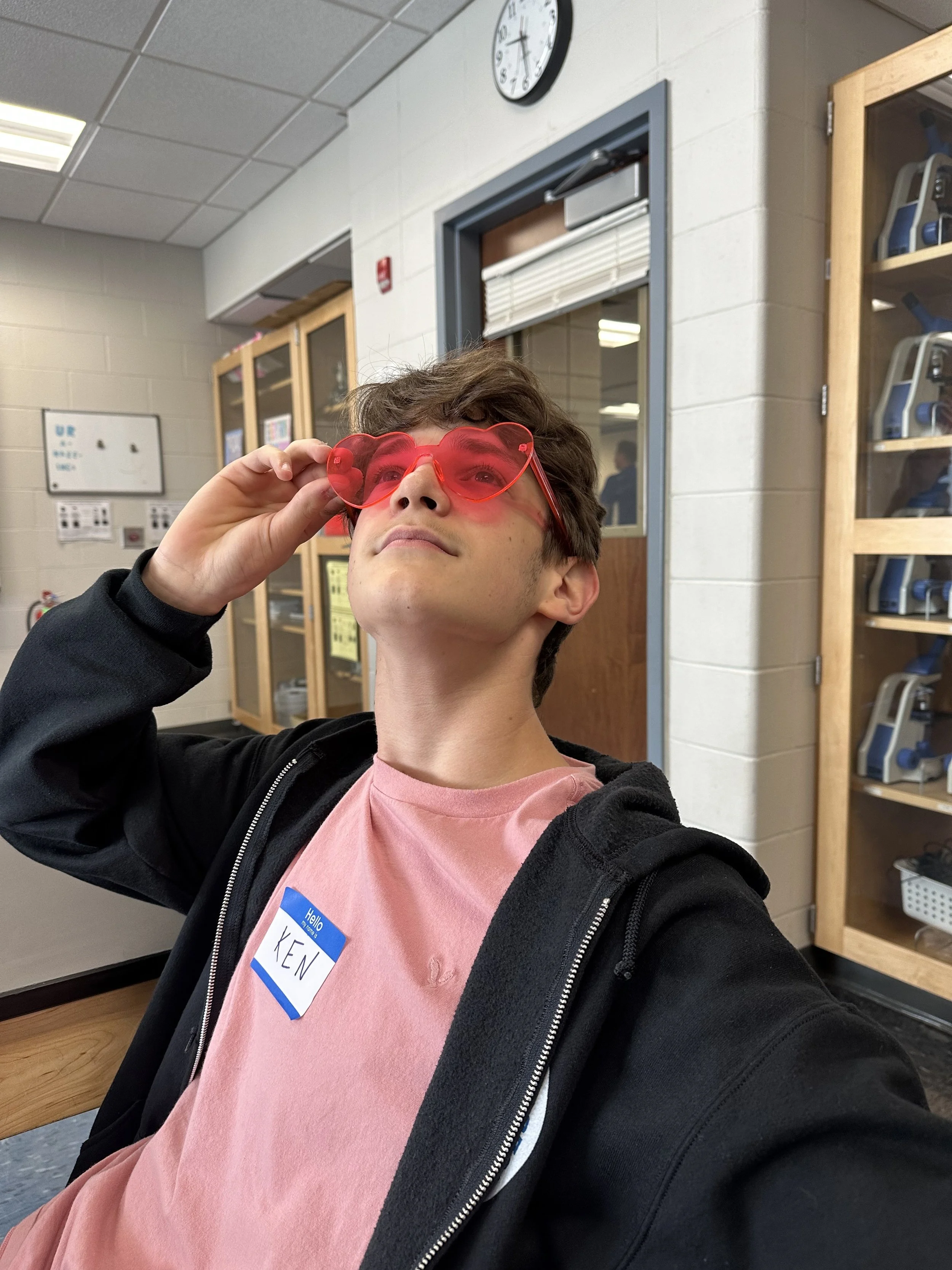 A young man with a nametag that reads 'KEN' wearing red heart-shaped sunglasses taking a selfie in a school or office setting with lockers and a clock in the background.