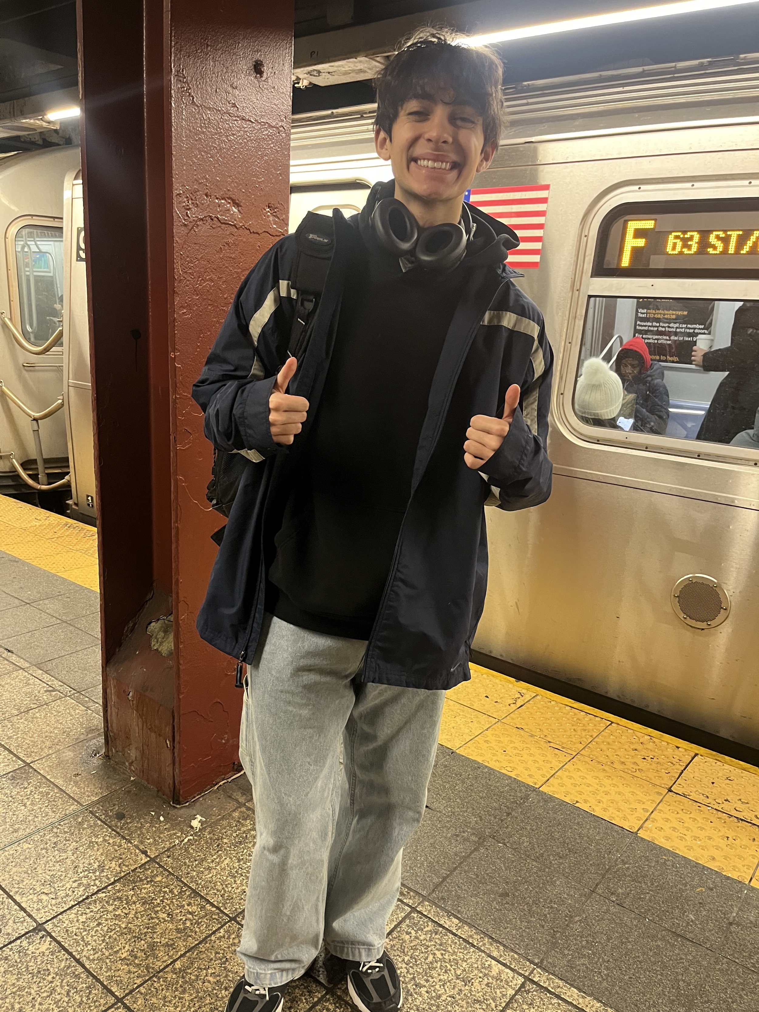 Young man with curly hair, smiling and giving two thumbs up, standing on a subway platform with a train in the background.