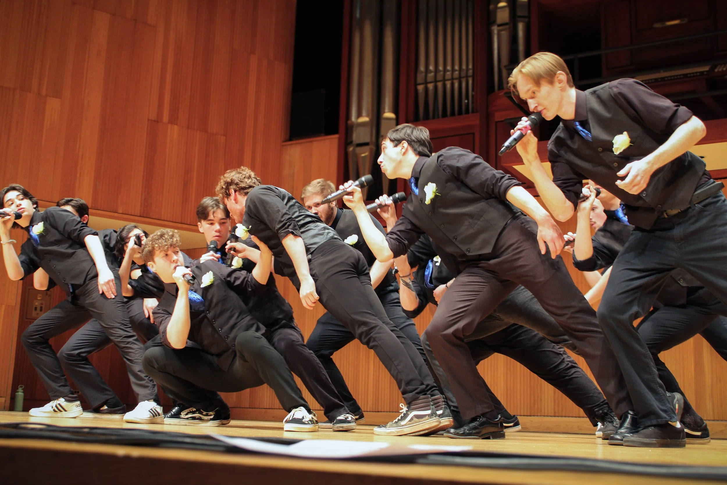 Group of young men performing a staged dance or musical act on a wooden stage, holding microphones, with a wooden backdrop and organ pipes in the background.