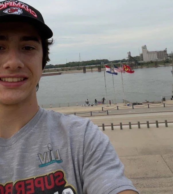 A young man wearing a gray shirt and a black cap taking a selfie near a body of water with three flags flying on poles, a walkway, and buildings in the background.