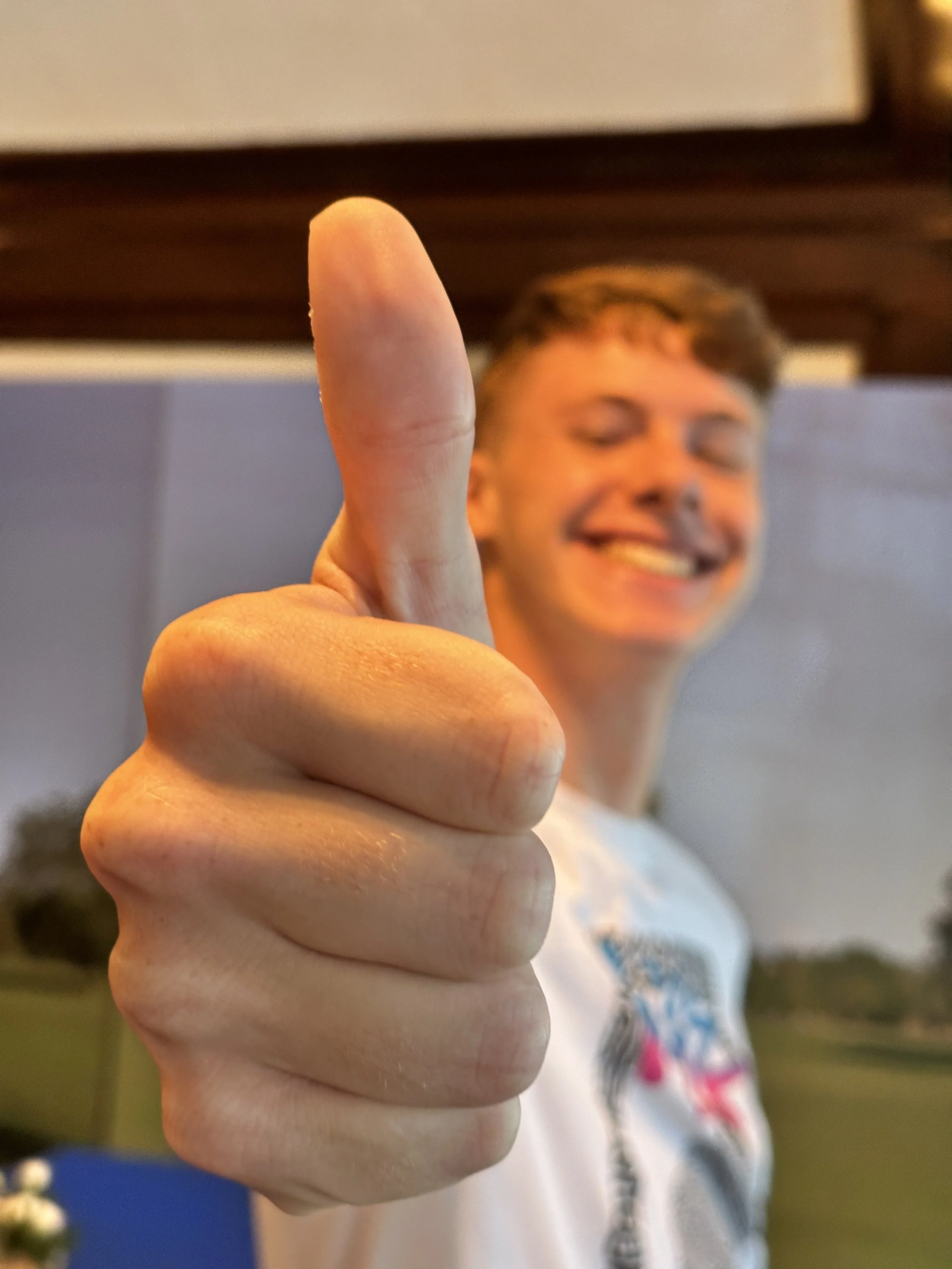 A young man smiling and giving a thumbs-up gesture, with the focus on his thumb in the foreground.