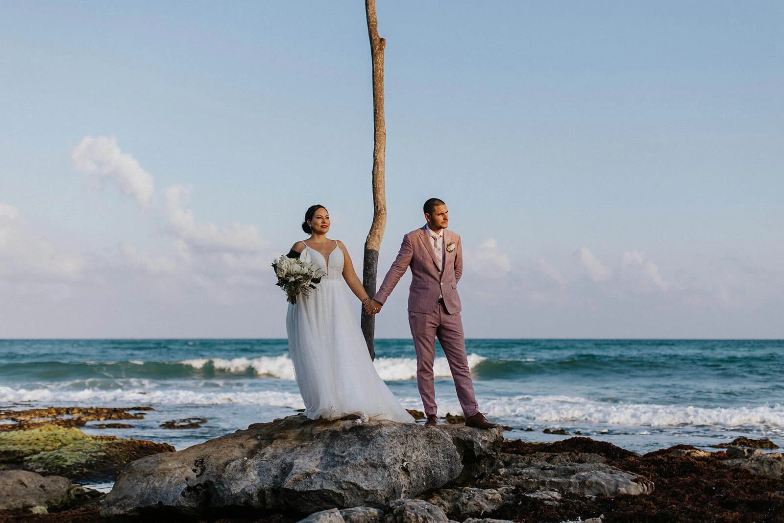 “Bride and groom holding hands on a rocky beach in the Riviera Maya, with ocean waves behind them during their wedding day.”