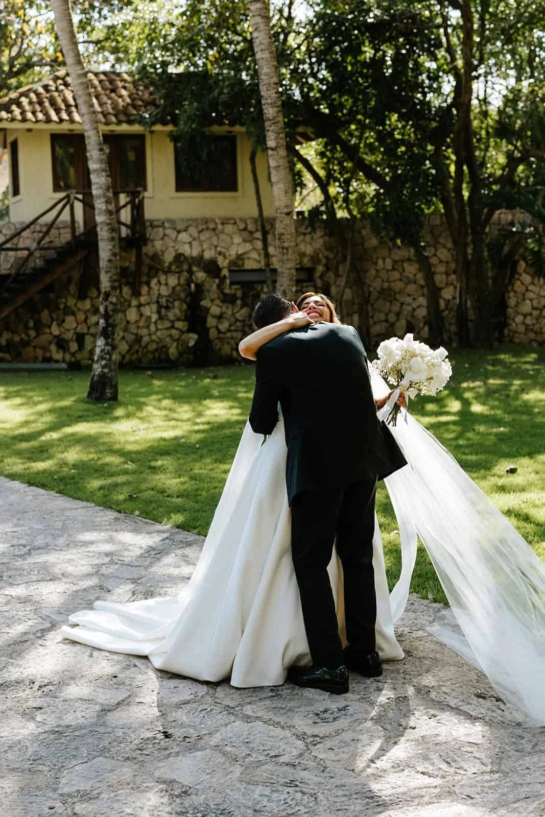  Bride and groom embracing after first look at Xcaret Park, groom holding bride, bride's white dress train flowing, palm trees and stone architecture in background, intimate emotional moment; 