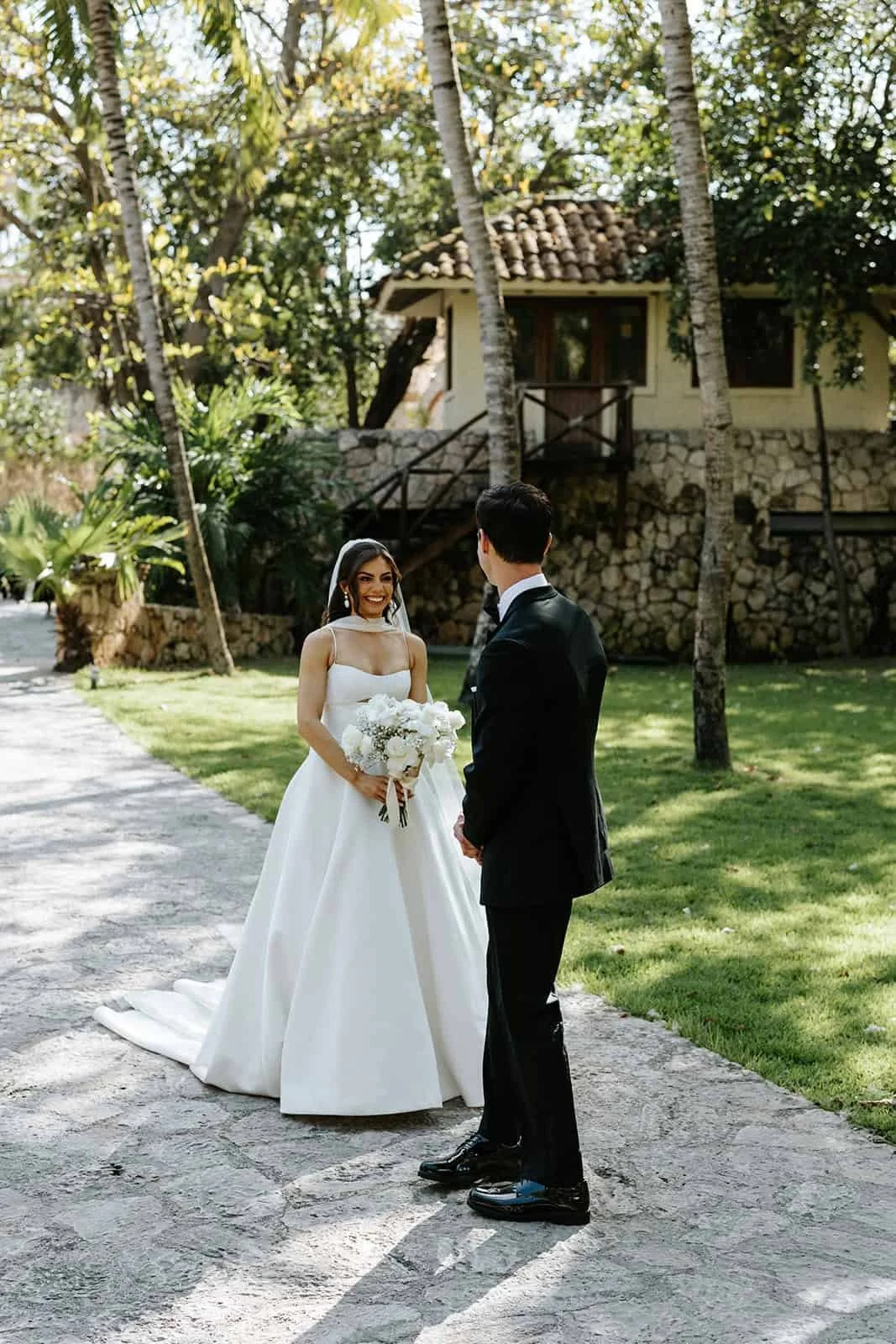  Bride and groom facing each other during first look at Xcaret Park, bride smiling, groom gazing at bride, palm trees and stone hacienda in background, bride holding white bouquet 