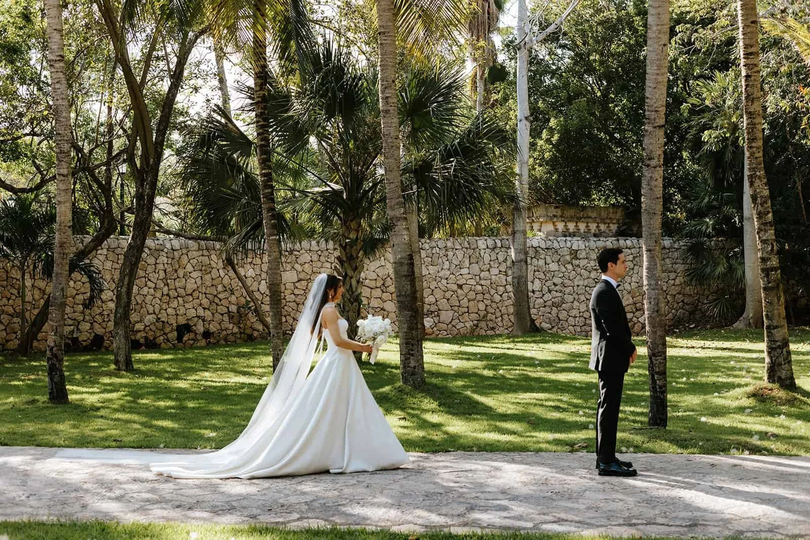  Bride and groom during first look at Xcaret Park — bride turns to see groom for first time, groom's back to camera, stone wall and palm trees framing the moment, white dress train flowing 