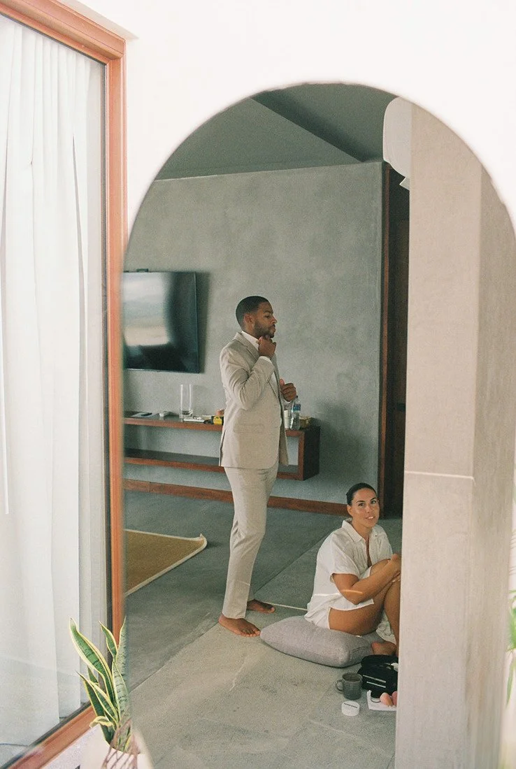 A reflection in a mirror shows a man in a beige suit standing and looking at his phone, while a woman in a white shirt is sitting on a cushion on the floor, looking up and smiling.