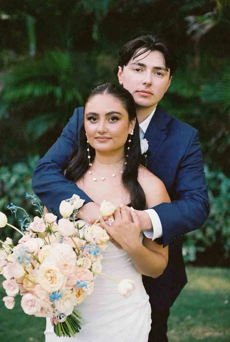 A newlywed couple stands outdoors, with the groom in a navy suit and the bride in a white wedding dress holding a bouquet of pastel-colored flowers.