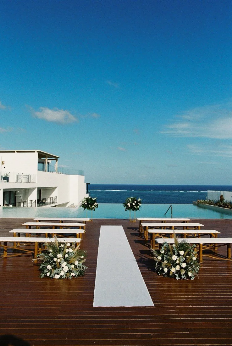 Outdoor wedding setup with white aisle runner, floral arrangements, benches, pool, ocean view, and modern white buildings under a clear blue sky.