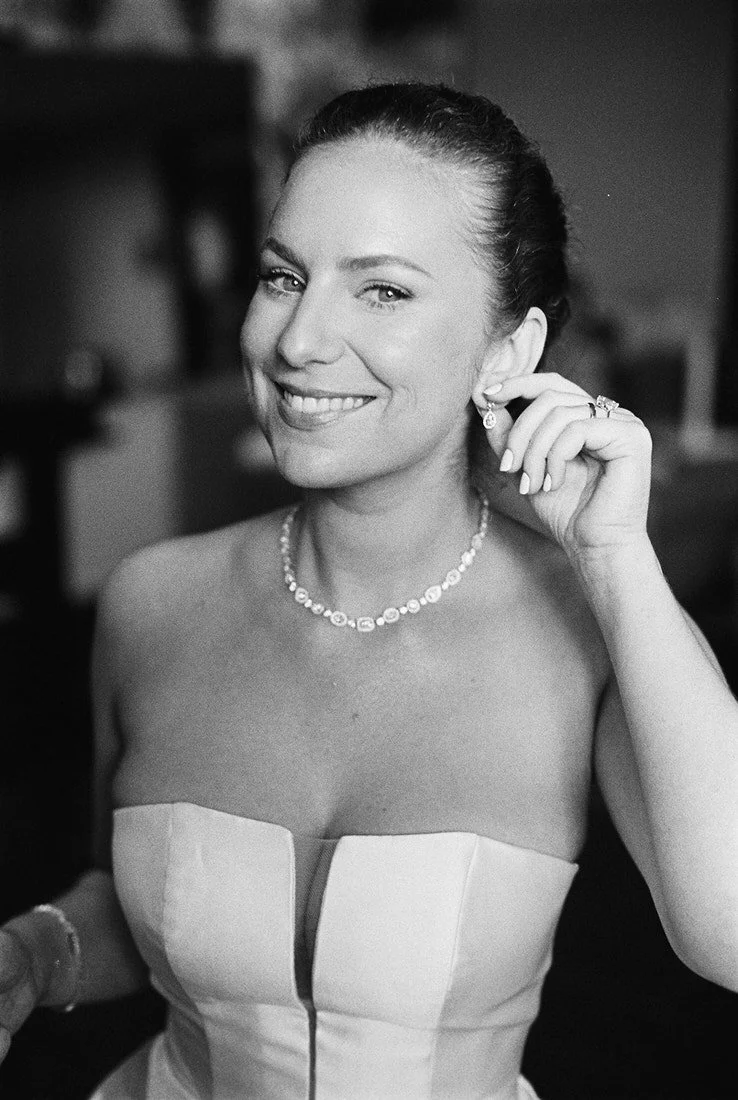 A smiling woman in a strapless dress, wearing jewelry including earrings, a necklace, and rings, adjusting her earring.